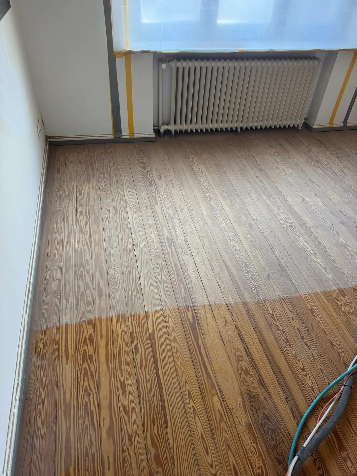 Dielenboden während der Arbeit: vorn noch die alte, dunkle Beschichtung, dahinter das bereits gestrahlte, helle Holz – Fenster abgedeckt, Wände abgeklebt